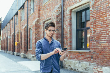 Young businessman uses phone outside office, happy and smiling man with smartphone