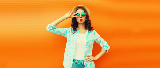 Portrait of beautiful young woman blowing her lips sending sweet air kiss posing wearing summer straw hat on orange background