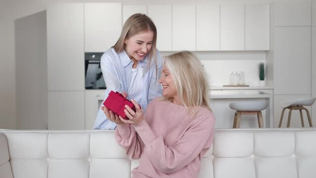 Loving adult daughter giving present to her senior mother. Young woman giving surprise bithday gift box, congratulating with hugs senior mom on mothers day on sofa at home