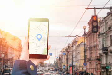 A woman's hand in a jacket holds a smartphone with an online map app. In the background is a city street with a green location icon.Light.Concept of Internet navigation and modern technologies