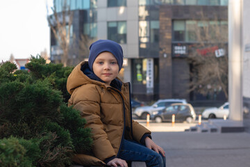 Fototapeta premium a boy in a jacket and hat sits on a bench in the city