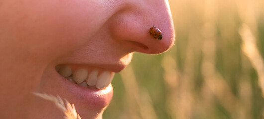 Young woman playing with a cute ladybug in the nature at sunset. Back to the nature concept. Ladybug is walking on the woman nose and she is smiling.