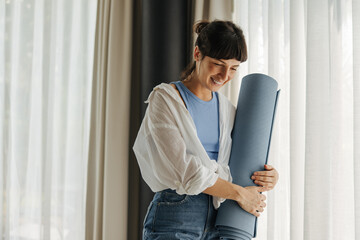 Beautiful young caucasian woman holding yoga mat in light room. Brunette wears casual clothes. Rest time concept. - obrazy, fototapety, plakaty