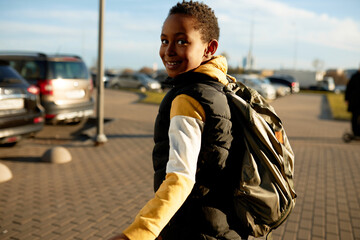 Rare view of african preteen kid with short afro haircut walking on sidewalk in city center, turning head to camera, carrying backpack on shoulders, going to school in sunny spring morning