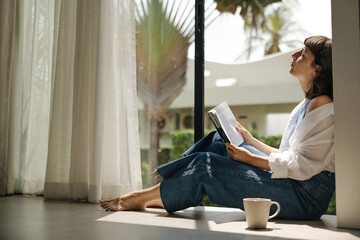 Calm young caucasian woman at home relaxing in living room reading book and drinking coffee. Brunette closed her eyes and basked in sun sitting near window. Relaxed lifestyle, concept
