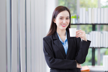 Asian business female who wears blue shirt and black suit holds coffee cup in her hand and smile happily at work office as background.