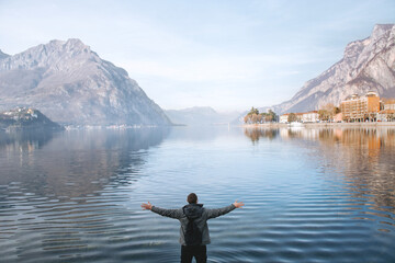 Blurred focus. Man traveler enjoys freedom with raised hands and backpack on the shores of Lake Lecco, Italy. Blue water, mountains and sky. Concept of tourist lifestyle, summer vacation outdoors.
