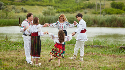 Happy family with kids in traditional romanian dress in a countryside, park. Father, mother, son and daughters dancing outside. - obrazy, fototapety, plakaty