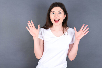 young beautiful Caucasian woman wearing white T-shirt over grey wall raising hands up, having eyes full of happiness rejoicing his great achievements. Achievement, success concept.