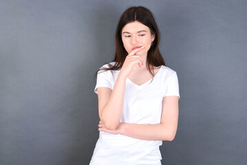 Shot of contemplative thoughtful young beautiful Caucasian woman wearing white T-shirt over grey wall keeps hand under chin, looks thoughtfully upwards.