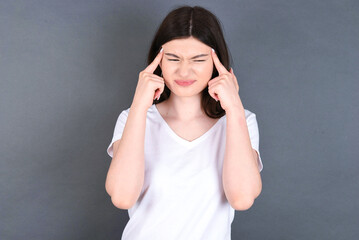 young beautiful Caucasian woman wearing white T-shirt over grey wall concentrating hard on an idea with a serious look, thinking with both index fingers pointing to forehead.