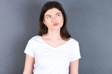 young beautiful Caucasian woman wearing white T-shirt over grey wall, looks pensively aside, plans actions after university, imagines what to do Thinks over about new project.