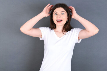 Fototapeta premium young beautiful Caucasian woman wearing white T-shirt over grey wall looks with excitement at camera, keeps hands raised over head, notices something unexpected, reacts on sudden news.