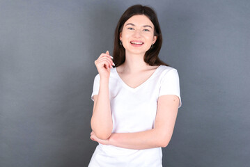 Fototapeta premium Cheerful young beautiful Caucasian woman wearing white T-shirt over studio grey wall with hand near face. Looking with glad expression at the camera after listening to good news. Confidence.