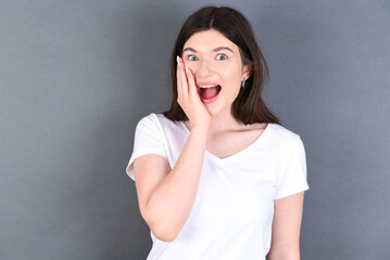 Shocked, astonished young beautiful Caucasian woman wearing white T-shirt ove looking surprised in full disbelief wide open mouth with hand near face. Positive emotion facial expression body language.