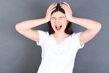 Shocked panic young beautiful Caucasian woman wearing white T-shirt over studio grey wall holding hands on head and screaming in despair and frustration.