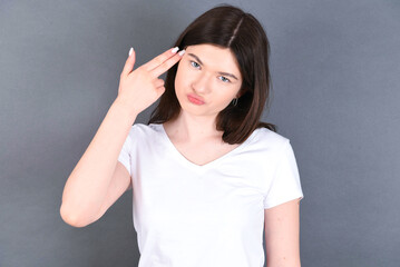 Fototapeta premium Unhappy young beautiful Caucasian woman wearing white T-shirt over studio grey wall imitates gun shoot makes suicide gesture keeps two fingers on temples.