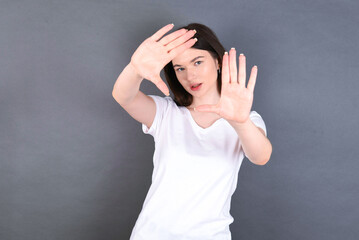 Portrait of smiling young beautiful Caucasian woman wearing white T-shirt over studio grey wall looking at camera and gesturing finger frame. Creativity and photography concept.