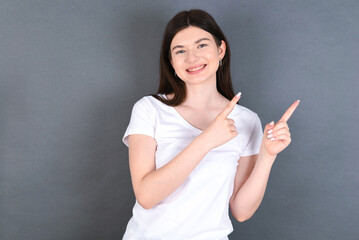 Positive young beautiful Caucasian woman wearing white T-shirt over studio grey wall with beaming smile pointing with two fingers and looking on empty copy space. Advertisement concept.