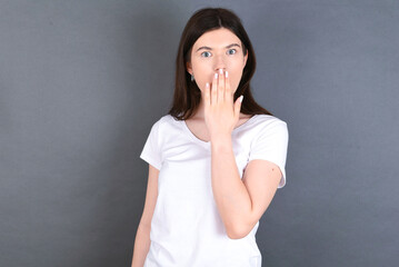 Oh! I think I said it! Close up portrait young beautiful Caucasian woman wearing white T-shirt over studio grey wall cover open mouth by hand palm, look at camera with big eyes.