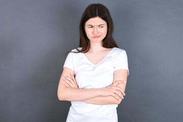 Displeased young beautiful Caucasian woman wearing white T-shirt over studio grey wall with bad attitude, arms crossed looking sideways. Negative human emotion facial expression feelings.