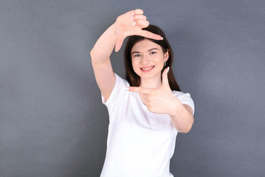 Positive young beautiful Caucasian woman wearing white T-shirt over studio grey wall with cheerful expression, has good mood, gestures finger frame actively at camera.