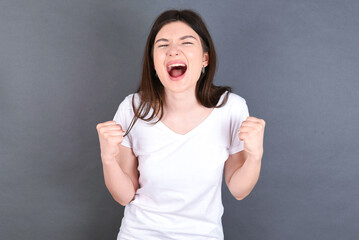 Portrait of young beautiful Caucasian woman wearing white T-shirt over studio grey  looks with excitement at camera, keeps hands raised over head, notices something unexpected reacts on sudden news.