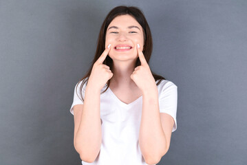 Strong healthy straight white teeth. Close up portrait of happy young beautiful Caucasian woman wearing white T-shirt over studio grey wall with beaming smile pointing on perfect clear white teeth.