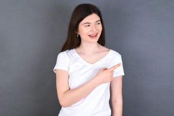 Fototapeta premium Happy cheerful smiling young beautiful Caucasian woman wearing white T-shirt over studio grey wall looking and pointing aside with hand. Copy space and advertisement concept.