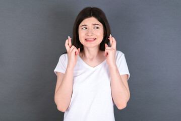 young beautiful Caucasian woman wearing white T-shirt over studio grey wall holding fingers crossed...