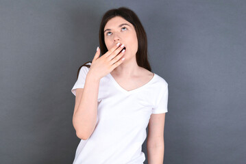 Sleepy young beautiful Caucasian woman wearing white T-shirt over studio grey wall yawning with messy hair, feeling tired after sleepless night, yawning, covering mouth with palm.