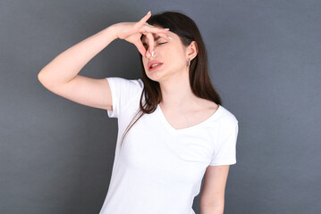 Displeased young beautiful Caucasian woman wearing white T-shirt over studio grey wall plugs nose as smells something stink and unpleasant, feels aversion, hates disgusting scent. - obrazy, fototapety, plakaty