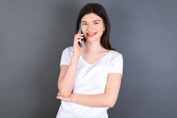 Portrait of a smiling young beautiful Caucasian woman wearing white T-shirt over studio grey wall talking on mobile phone. Business, confidence and communication concept.