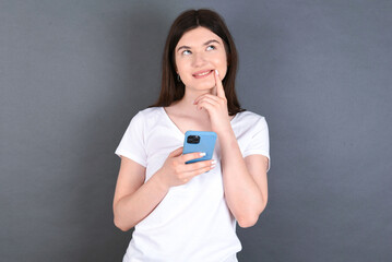 Image of a thinking dreaming young beautiful Caucasian woman wearing white T-shirt over studio grey wall using mobile phone and holding hand on face. Taking decisions and social media concept.