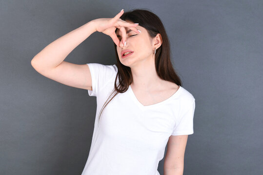 Displeased Young Beautiful Caucasian Woman Wearing White T-shirt Over Studio Grey Wall Plugs Nose As Smells Something Stink And Unpleasant, Feels Aversion, Hates Disgusting Scent.