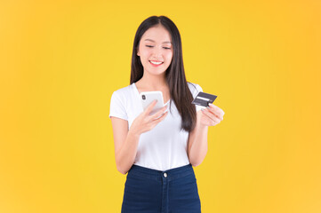 Naklejka premium pretty asian woman smile showing, presenting credit card for payment or paying online in holding mobile phone payment credit card for confident money and financial concepts yellow background.