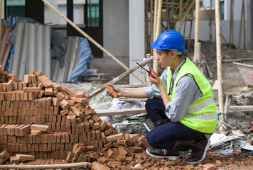 Asian contractor with safety helmet, reflective vest and rubber boots checking quality of brick...