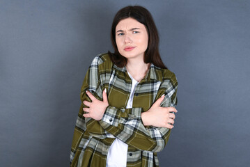 young beautiful Caucasian woman wearing plaid shirt over studio grey wall shaking and freezing for winter cold with sad and shock expression on face.