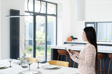 asian woman controlling smart home functions through tablet at modern home, looking at ceiling lamp