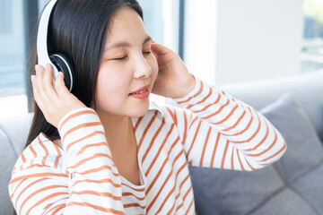 portrait of smiling young asian woman with closed eyes listening music in wireless headphone at home