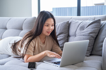 beautiful young asian woman in casual cozy clothes using laptop sitting on couch at modern apartment