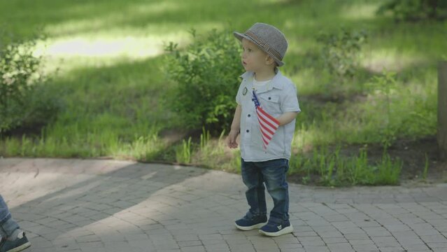 cute caucasian toddler child kid holding American Flag outdoors, baby in grey hat, blue jeans standing in green park, watching. national US flag symbol celebrating Memorial Day, Veterans Day, 4th July