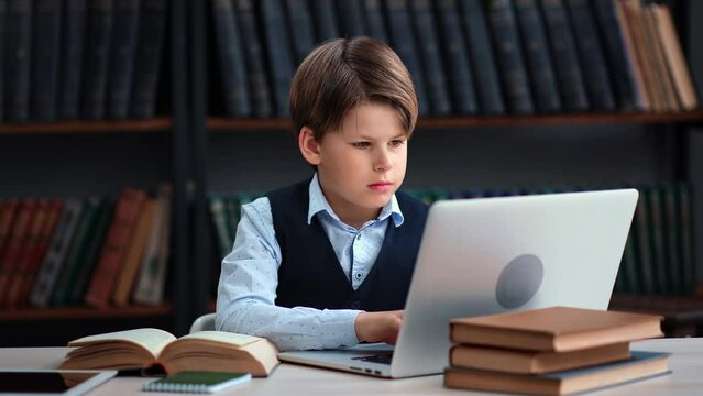 Modern male teen kid pupil e learning homework paper book chatting laptop at library desk