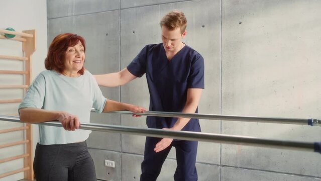 Physical Therapy Center: Portrait of Strong Senior Woman with Leg Injury Successfully Walks Holding Parallel Bars. Physiotherapist, Rehabilitation Doctor, Trauma Prevention Therapy Hospital.