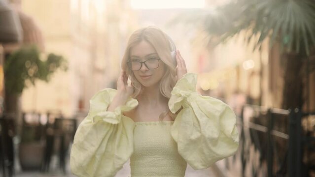 Closeup face of beautiful young blond girl with long hair and eyeglasses. Slow motion of smiling young girl listening to music in headphones at the city street. 
