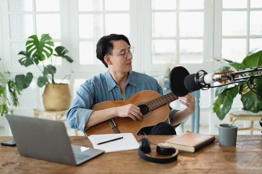Young Asian artist musician man sitting in living room singing a song while work and playing guitar at home studio