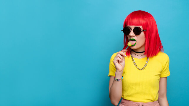 A bright and funny young woman with red hair licks a lollipop. Studio colorful picture of a pin-up style woman with a lollipop on a stick, on a blue background. A place for your text.