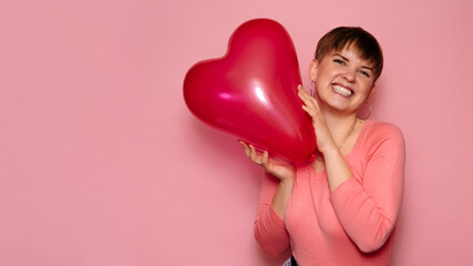 Portrait of a bright young woman with a red heart-shaped balloon on a pink background. A place for your text.