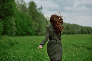 Fototapeta premium a woman in a long raincoat runs across a field in tall green grass in cloudy weather in spring