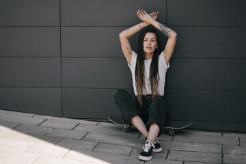 Young woman with braids and tattoos wearing casual clothes standing with skateboard on city street.